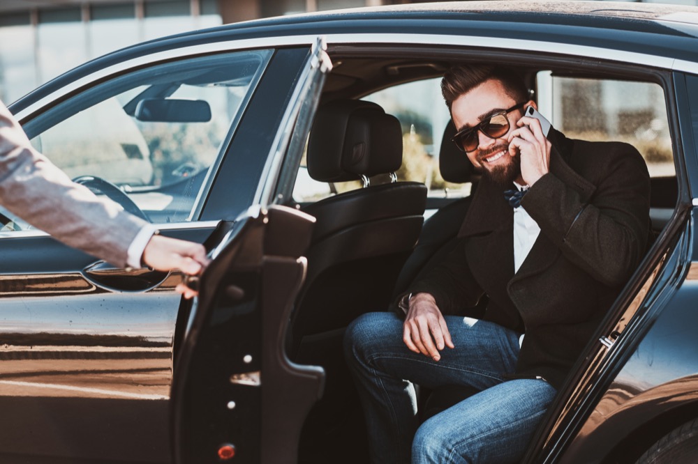 Busy smiling businessman in sunglasses is talking by smartphone while his elegant assistant is opening door for him.