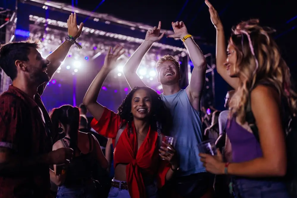 A young crowd of multiethnic friends are dancing at a concert having beers and holding their hands up in their casual clothing. 
