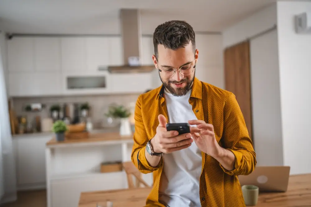man in glasses standing in his home kitchen using phone to book online
