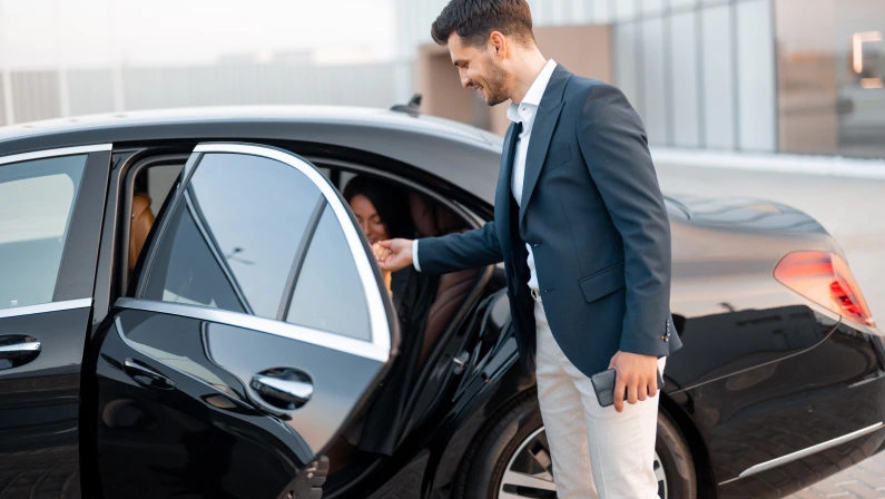 Businessman helps gently a woman to get out of a car, arrived by luxury vehicle for some event at evening. Concept of transportation and business lifestyle